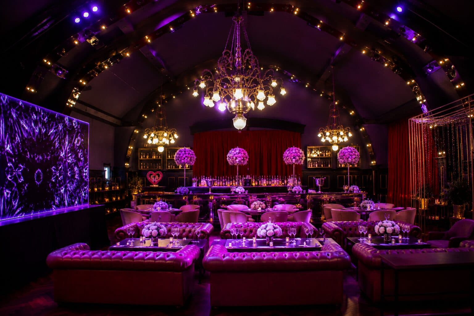 El salón ambientado en violeta con sillones Chester alrededor de mesas bajas con flores, arañas encendidas y una pantalla gigante sobre la pared.