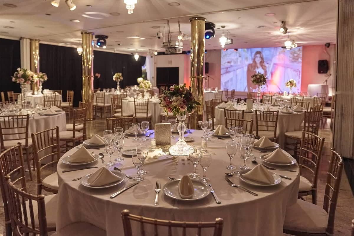 El salón montado con mesas redondas de mantel blanco y sillas doradas tipo tiffany, centros de flores altos, columnas doradas espejadas y al fondo una pantalla grande proyectando debajo de los equipos de luces colgados del techo.