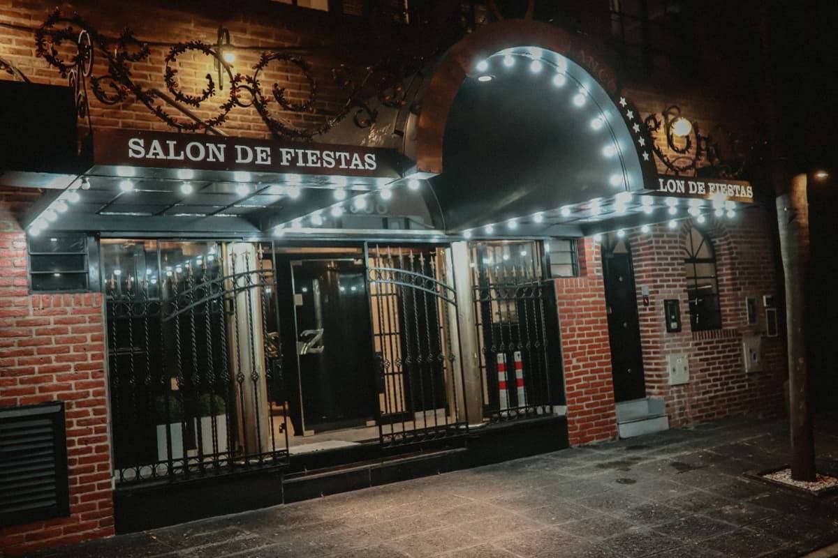 La fachada de ladrillo a la noche, con la marquesina de lamparitas que dice Salón de Fiestas, el arco de luces sobre la entrada y las rejas negras dando a la vereda.