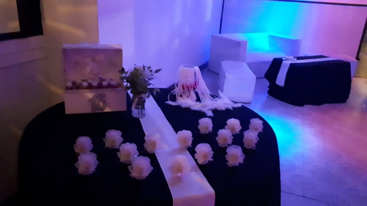 La mesa de recepción vestida de negro con rosas blancas y un cofre de madera, y al fondo un living blanco bajo luces azules y violetas.