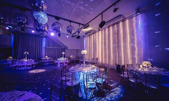 El salón armado para una cena, con mesas redondas de mantel claro, sillas Chiavari, centros de flores altos, bolas de espejo en el techo y una cortina de luces al fondo.