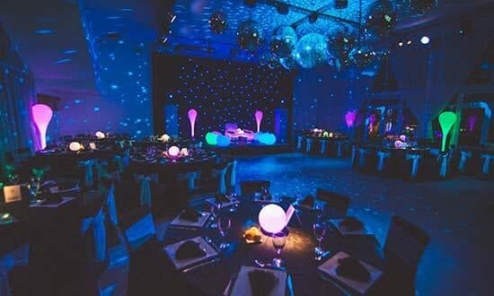 El salón con la iluminación de fiesta encendida en azul, con lámparas de colores repartidas entre las mesas, bolas de espejo y un telón de luces detrás de la pista.