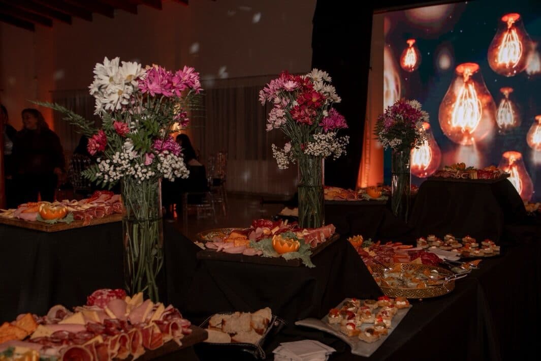 La mesa de recepción montada sobre manteles negros, con tablas de fiambres y quesos, bandejas de canapés y tres floreros altos de margaritas blancas y claveles rosados, y al fondo una pantalla de gran formato encendida con farolas de luz cálida.