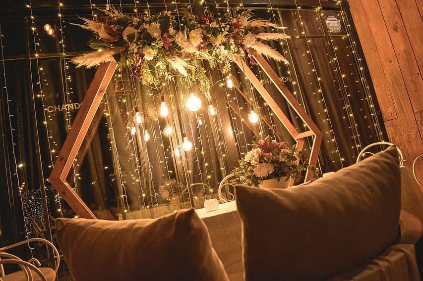 Un arco hexagonal de madera con flores y pampas arriba, colgado de guirnaldas de luces y lámparas, con sillones de almohadones adelante.