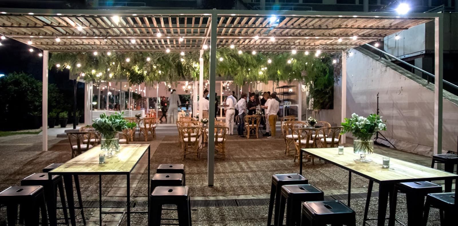 La pérgola de listones de madera iluminada de noche con guirnaldas de luces y helechos colgando, mesas altas con banquetas adelante y el salón vidriado al fondo.