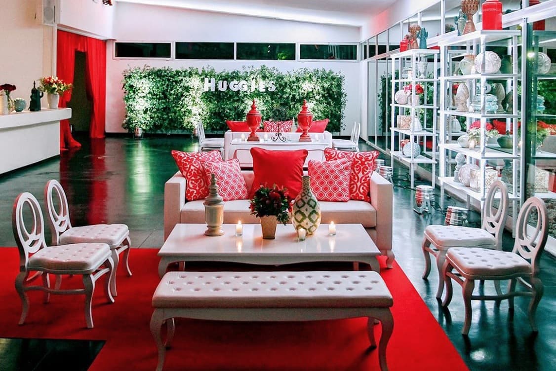 El salón interior con piso oscuro espejado, sillones y mesas bajas blancas con almohadones rojos sobre una alfombra roja, y un muro de follaje con letras luminosas al fondo.