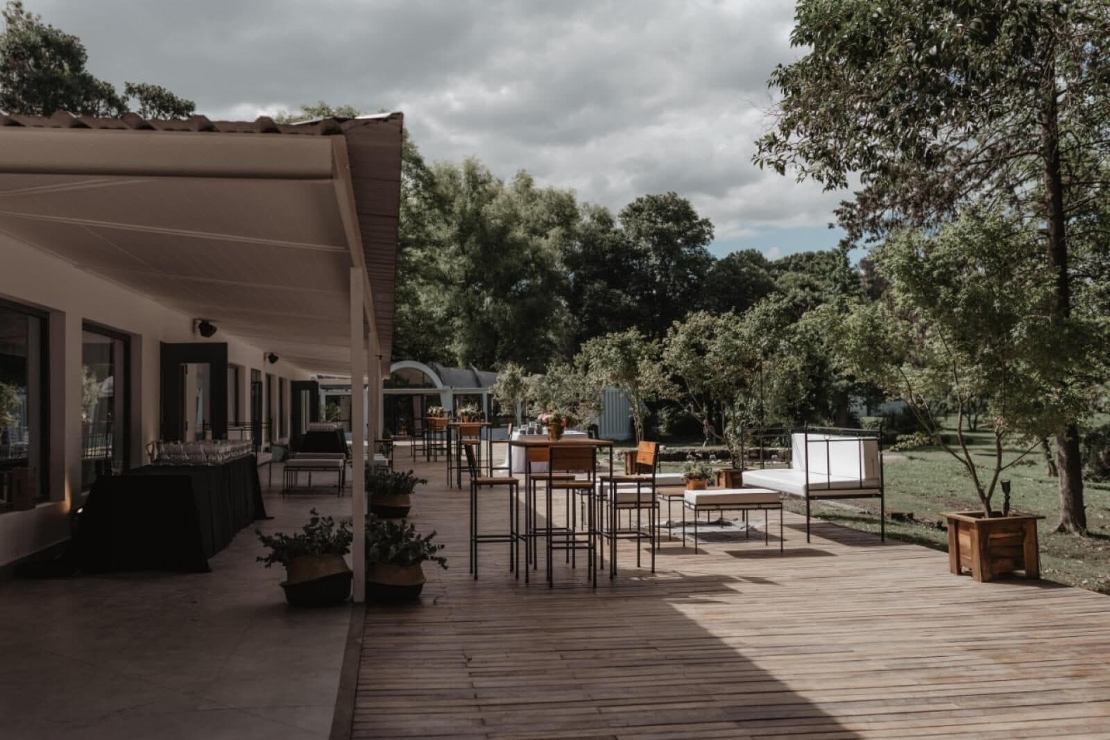 El deck de madera bajo el alero del salón, con mesas altas y banquetas de hierro, sillones blancos de exterior, macetones de madera y una barra con mantel negro contra los ventanales, y el parque arbolado al costado.