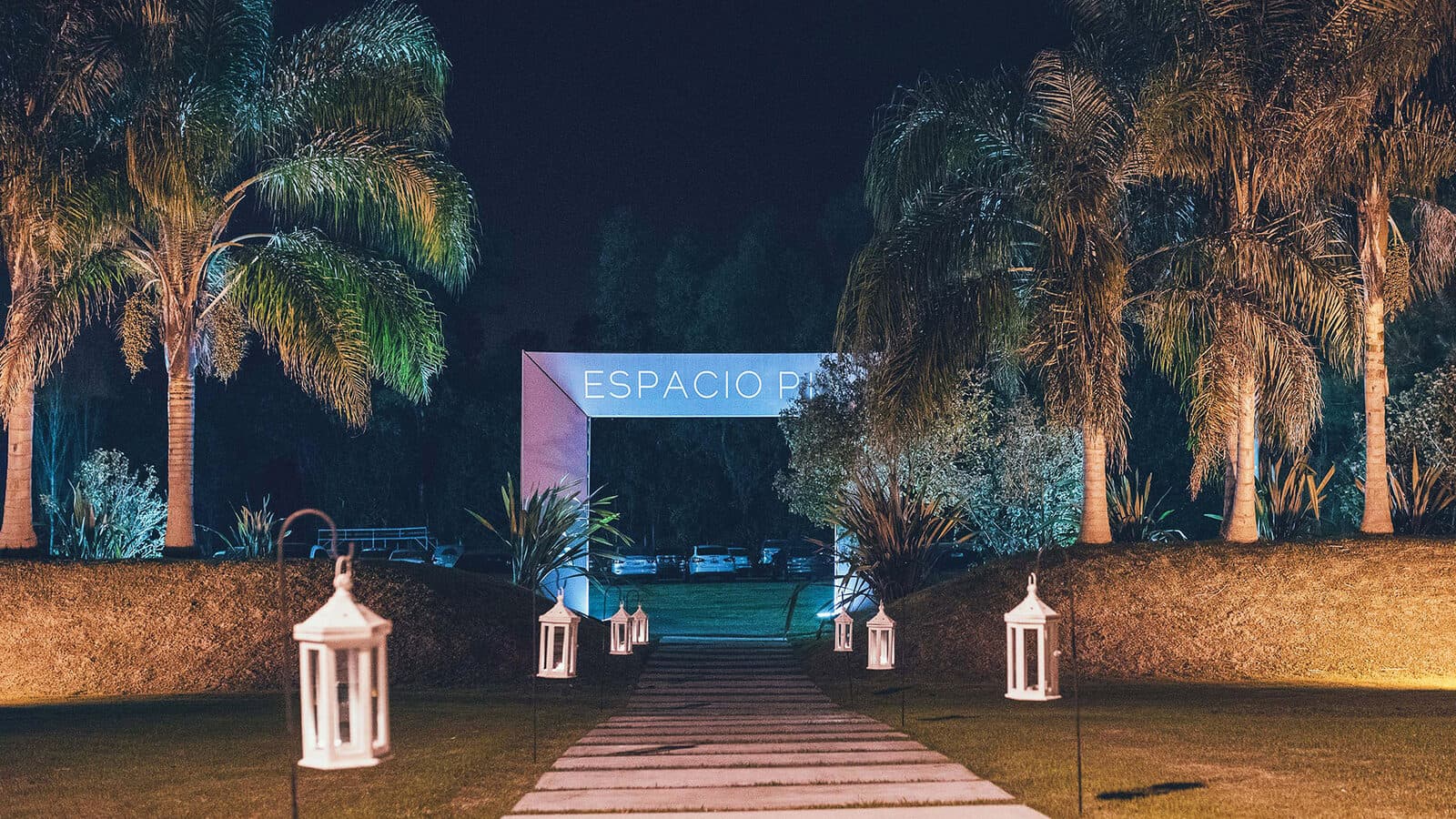 La entrada de noche, con el camino iluminado entre faroles blancos altos, palmeras a los dos lados y el cartel del lugar encendido al fondo.
