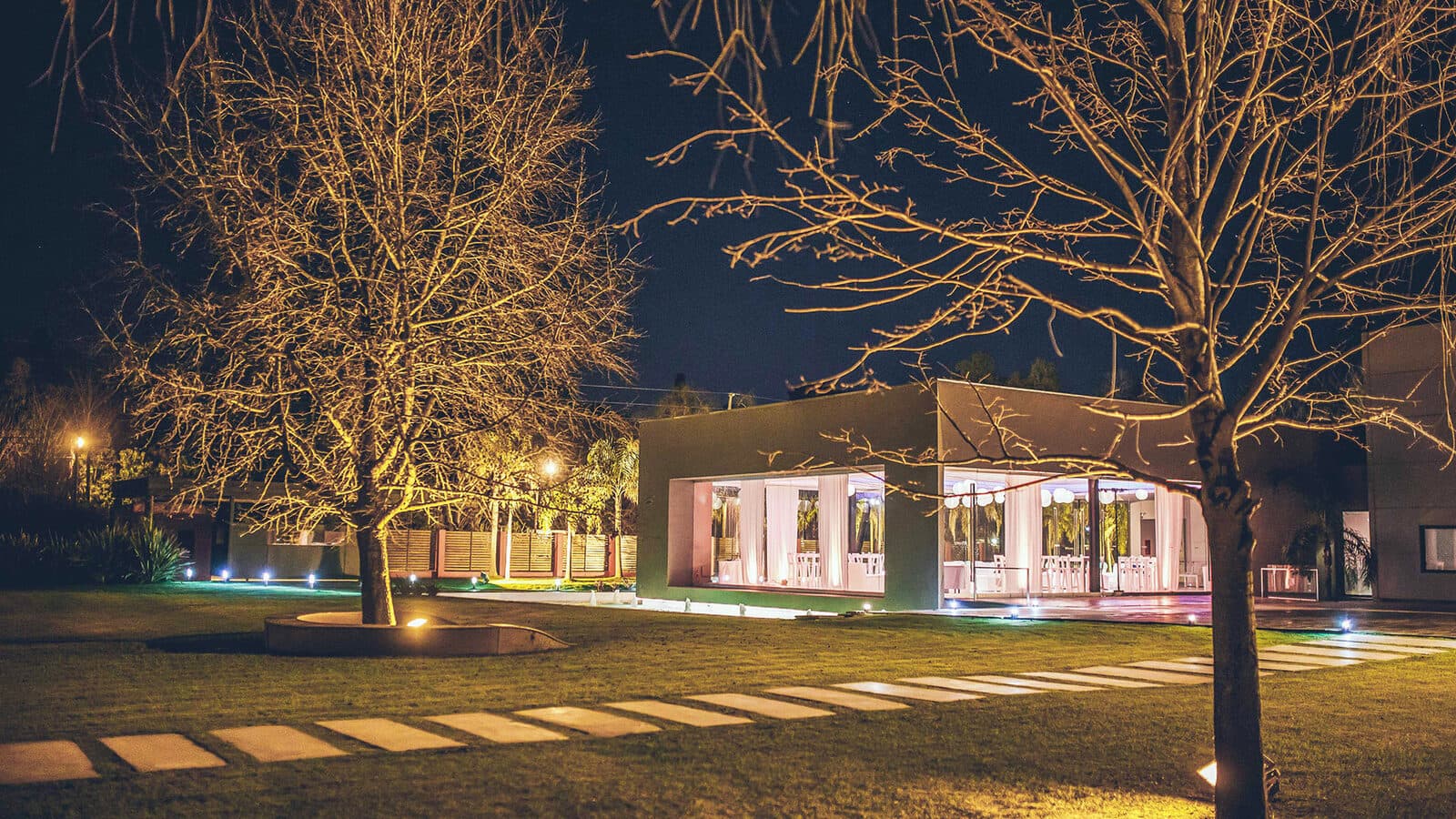 El salón de noche visto desde el parque, con los ventanales encendidos, luces bajas sobre el césped y árboles sin hojas recortados contra el cielo.