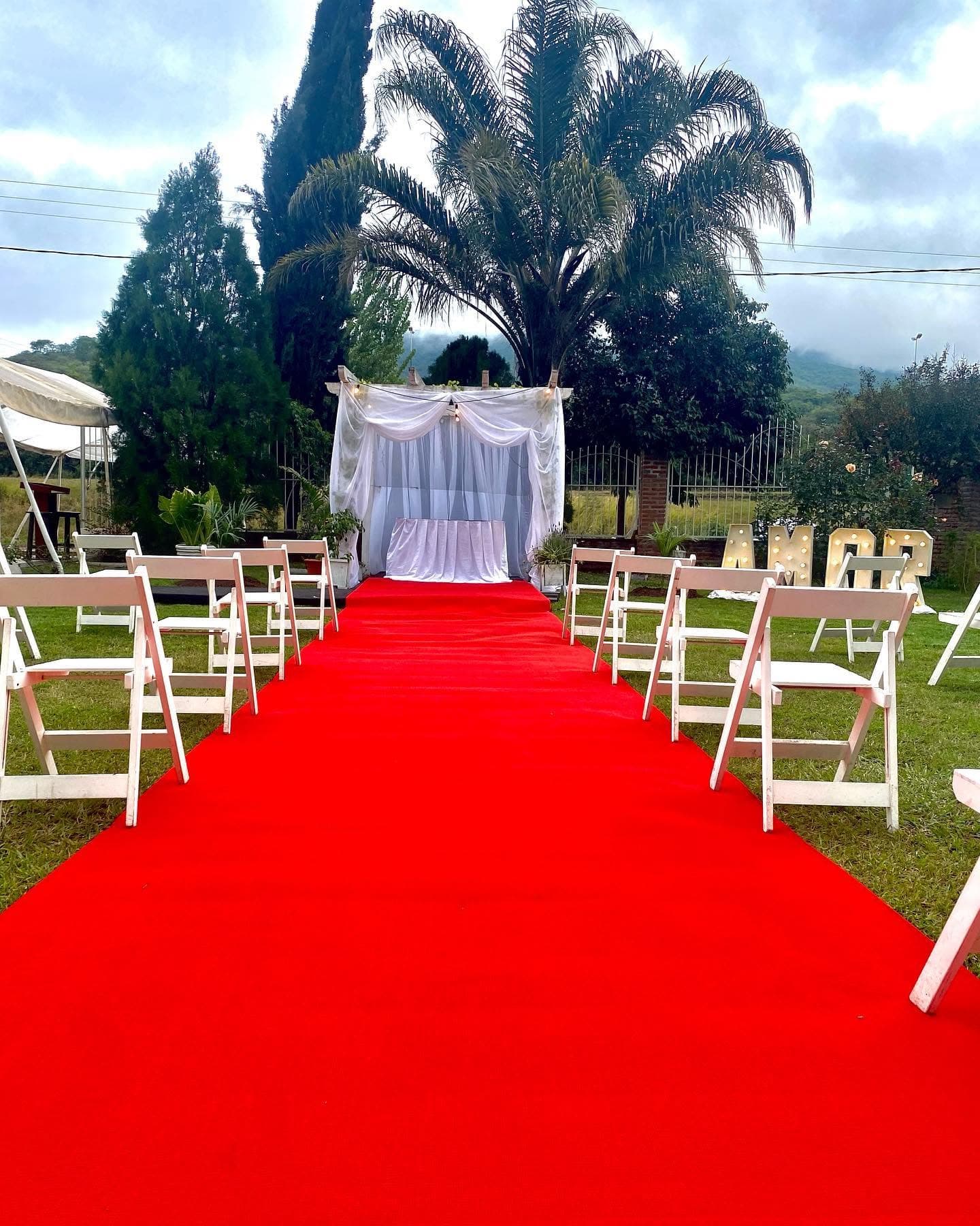 La ceremonia montada en el parque: una alfombra roja entre dos hileras de sillas blancas hasta un arco con cortinados, con las palmeras y los cerros detrás y unas letras AMOR iluminadas a un costado.