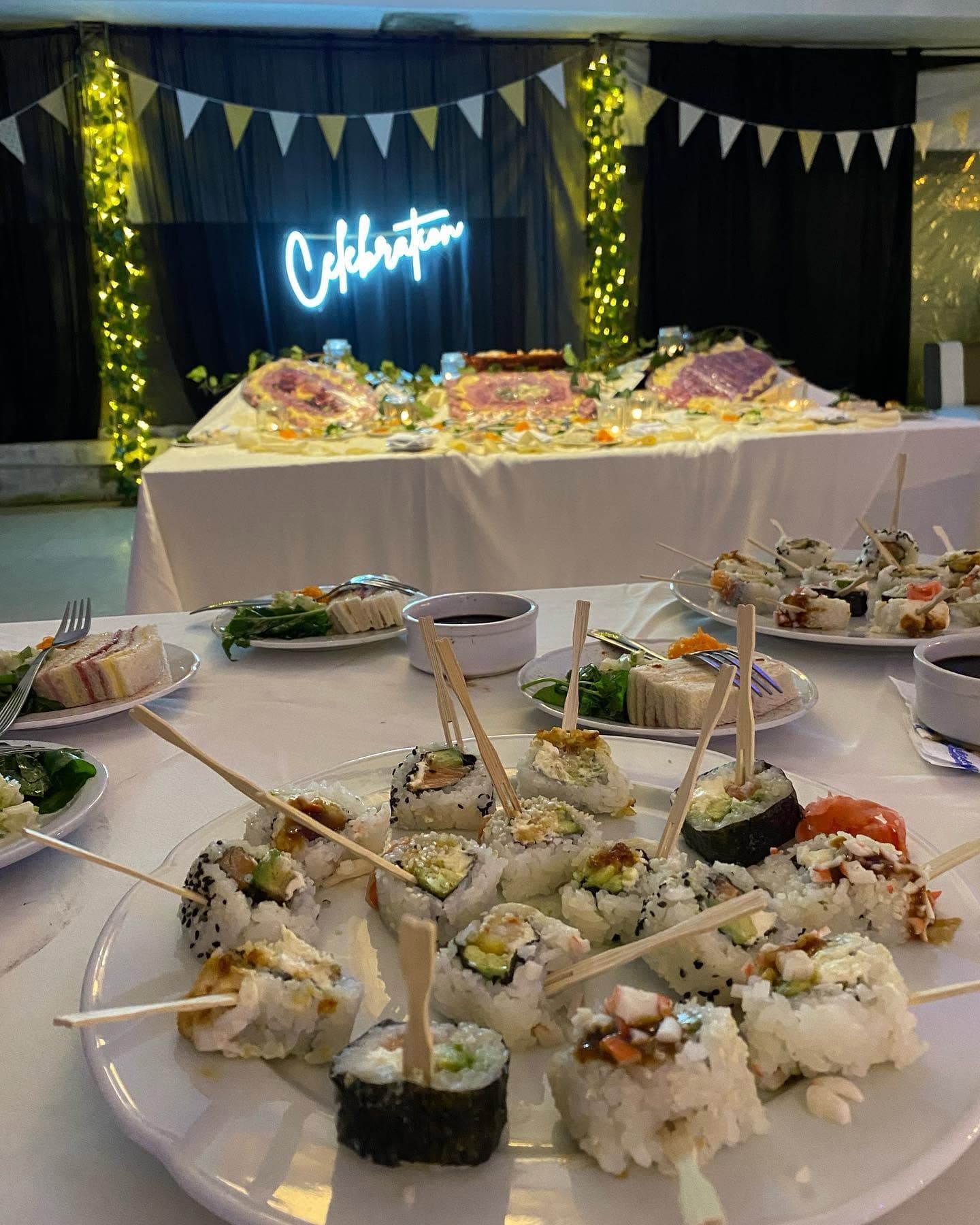 Una fuente de sushi en primer plano sobre la mesa tendida, y al fondo la mesa principal con un cartel de neón que dice Celebration entre guirnaldas de luces.