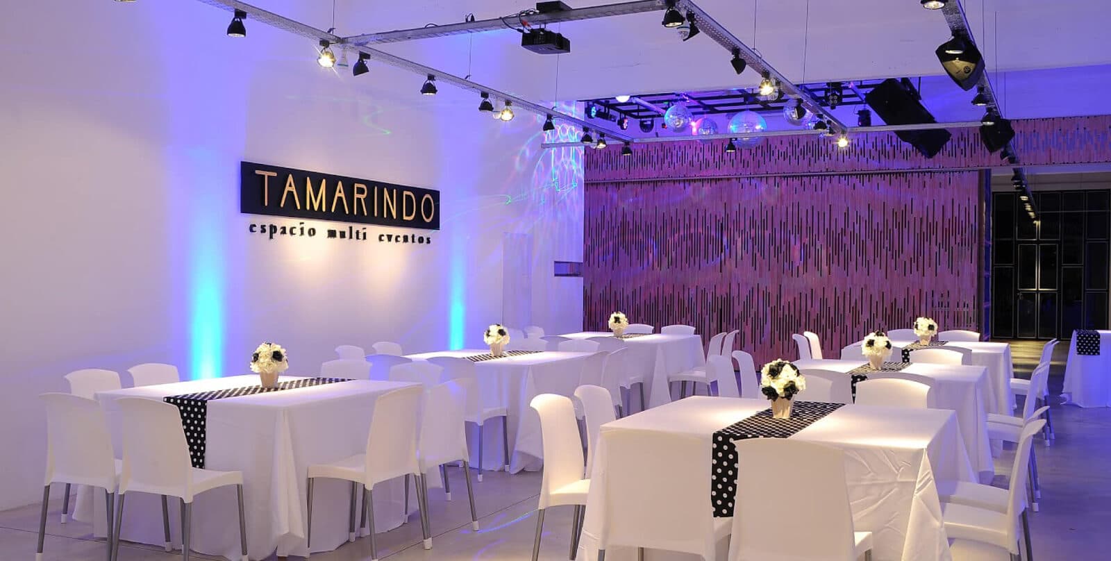 El salón armado con mesas de mantel blanco, caminos de mesa a lunares y centros de flores, sillas blancas, el cartel del salón sobre la pared y una cortina de flecos violeta al fondo.