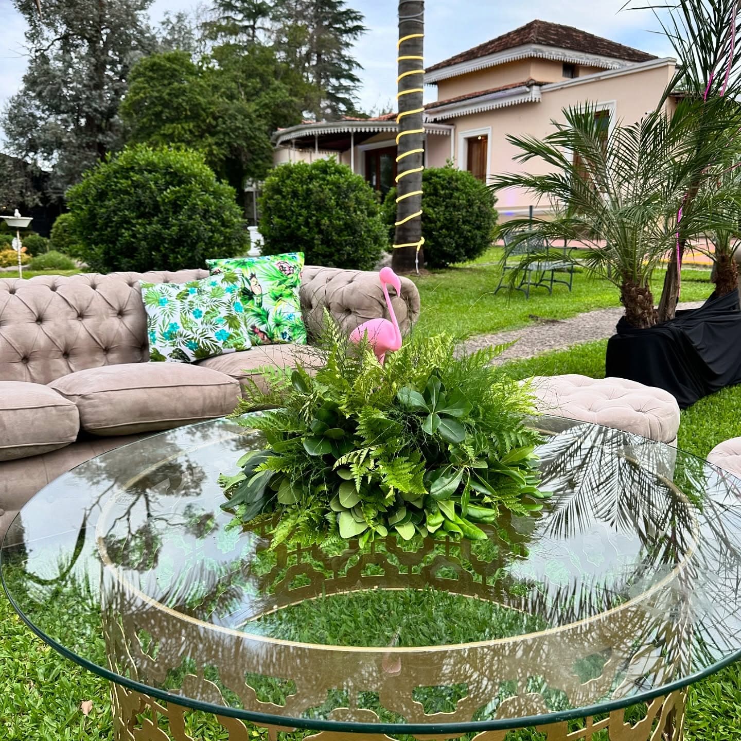 Un living de sillones sobre el césped con una mesa ratona de vidrio y un centro de helechos, y detrás la casona de techo de tejas entre las palmeras.
