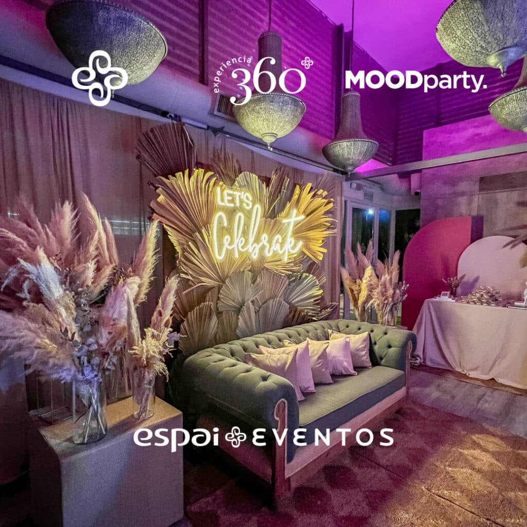 El living armado con un sillón capitoné verde, almohadones rosados, plumas de pampa y un cartel de neón que dice Let's Celebrate sobre un fondo de hojas de palma secas.