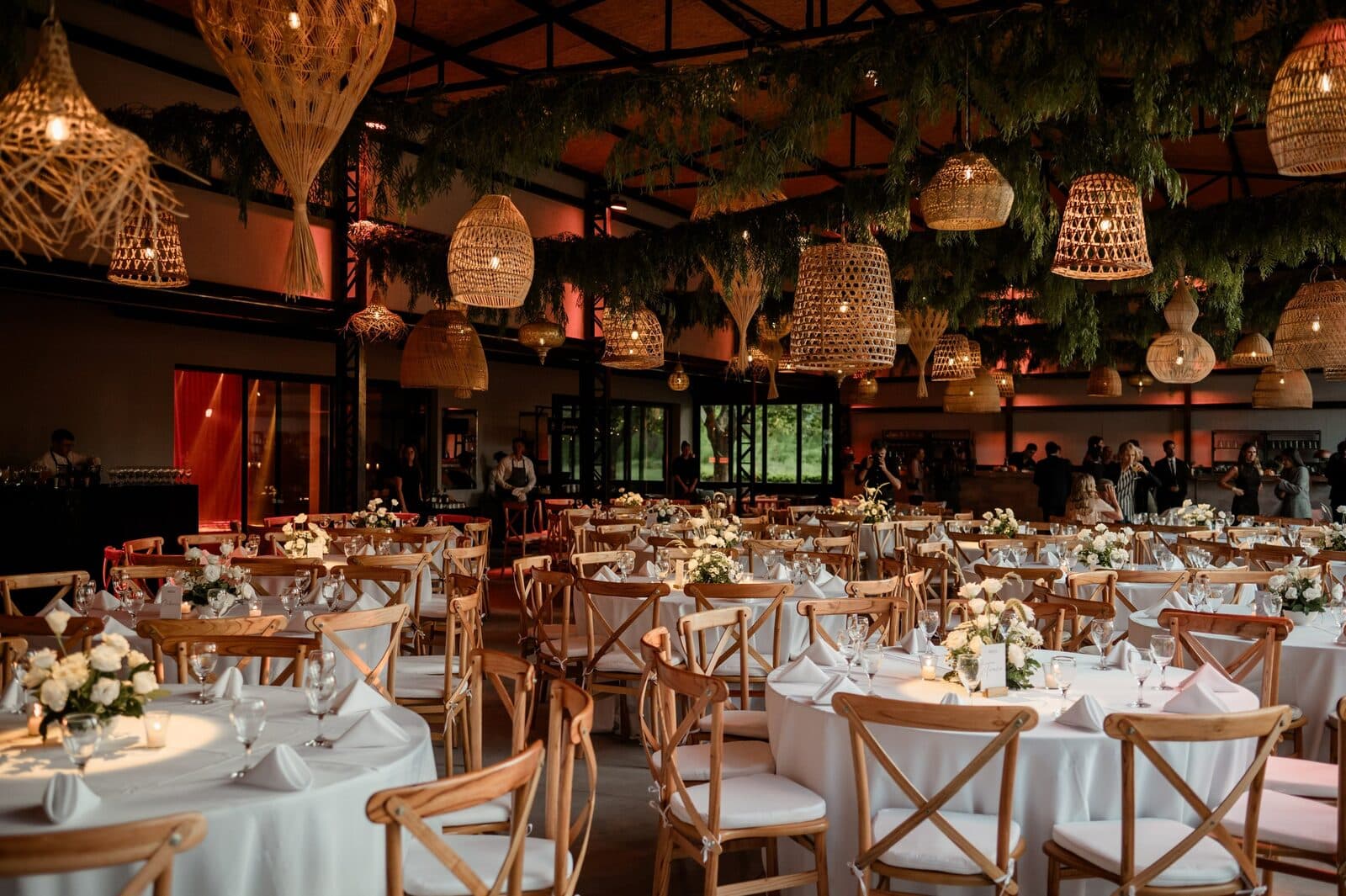 El salón techado con mesas redondas vestidas de blanco, sillas de madera clara y una hilera de lámparas de mimbre colgando entre plantas.