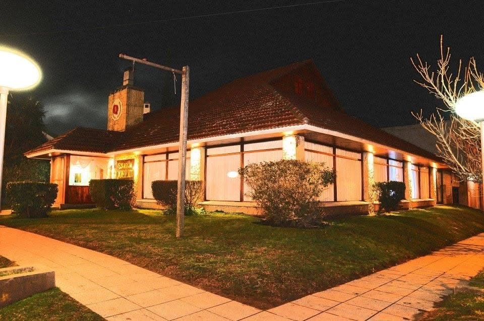 La casa de techo de tejas iluminada de noche, con los ventanales encendidos y el parque adelante.