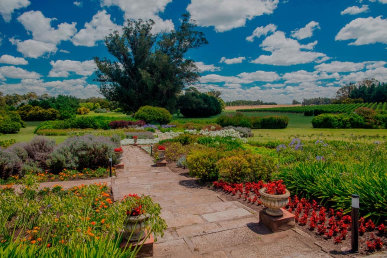 El jardín de la estancia con el camino de lajas entre los canteros floridos y el campo abierto al fondo.