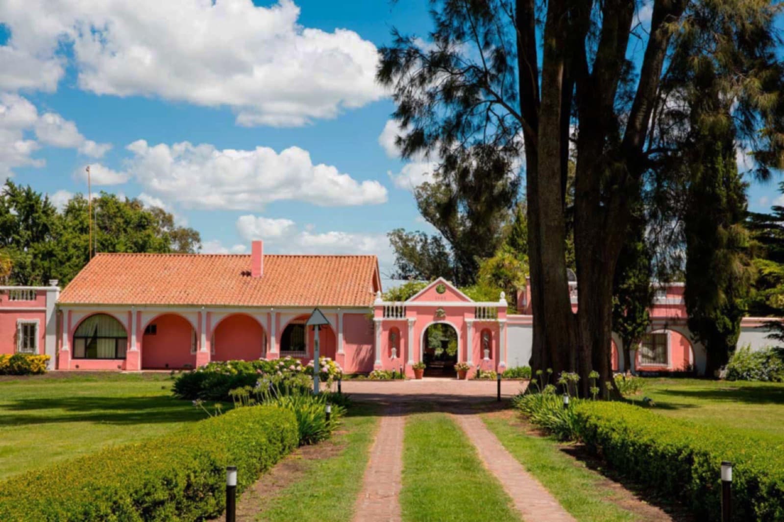 El casco rosado de la estancia visto desde el camino de entrada, con los cipreses altos a un costado y el parque recortado adelante.