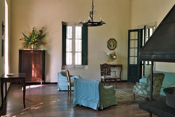 Un salón interior de paredes claras y techo alto, con araña de hierro con velas, piso de tablones de madera, sillones tapizados en celeste y verde, una cómoda antigua con un arreglo de flores y la ventana de postigos verdes que entra el sol.