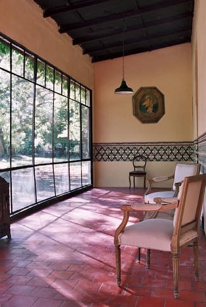 La galería vidriada con piso de baldosas coloradas y techo de vigas de madera, una lámpara colgante de campana verde, un cuadro antiguo sobre la pared con guarda calada, sillas y una mesita redonda, y el parque arbolado detrás del ventanal.