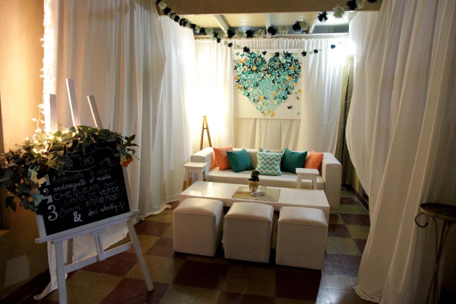 El sector de living con sillones blancos, almohadones turquesa y un corazón de mariposas de papel en la pared, entre cortinados claros.