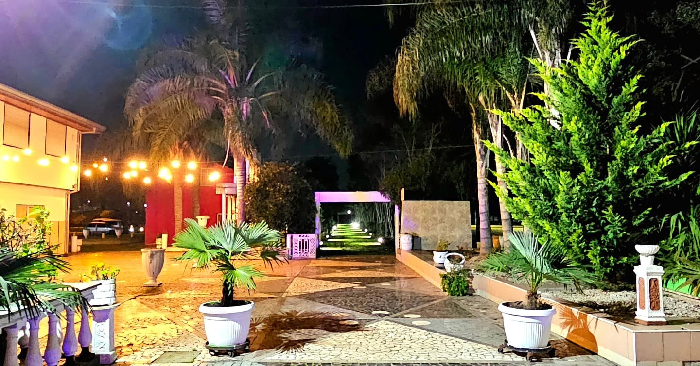 El parque de noche con palmeras iluminadas, macetas blancas y un patio de mosaicos, con la galería del salón encendida al fondo.