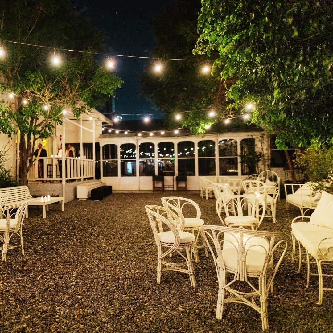 El patio al aire libre de noche, con piso de piedra, sillones y sillas de mimbre blanco, guirnaldas de luces entre los árboles y la galería vidriada al fondo.