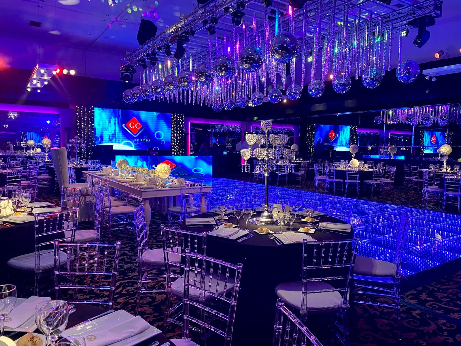 El salón entero en luz azul, con las mesas puestas, sillas transparentes, candelabros altos de centro de mesa y pantallas encendidas sobre la pista.
