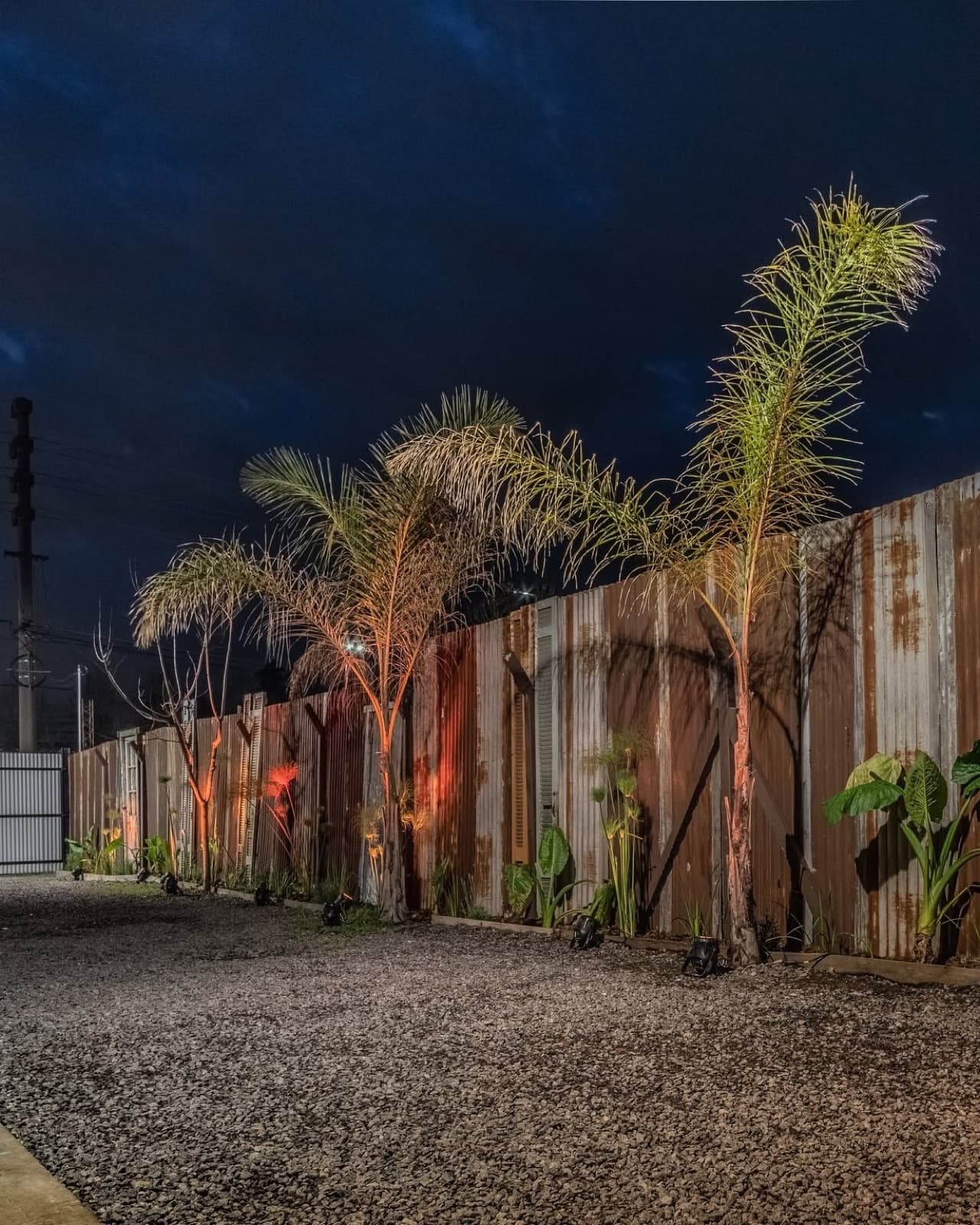 El patio de entrada de noche, con la pared de chapa oxidada iluminada desde abajo, palmeras y plantas contra el muro y el suelo de piedra partida.