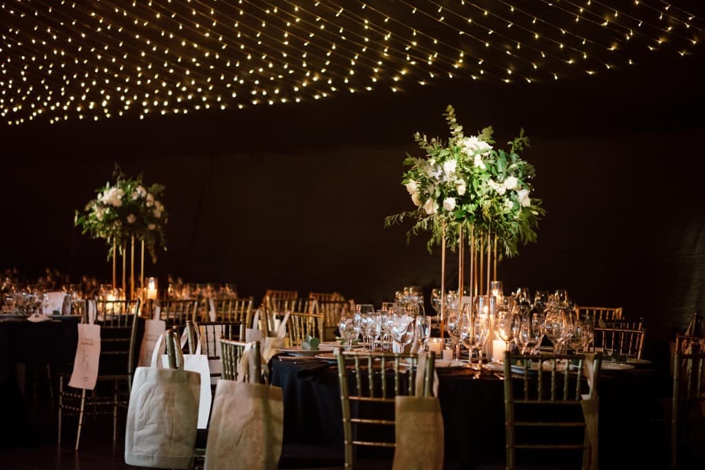 Las mesas vestidas de negro con centros altos de flores blancas y verdes, las sillas de madera clara y un cielo de guirnaldas de luces sobre la carpa.
