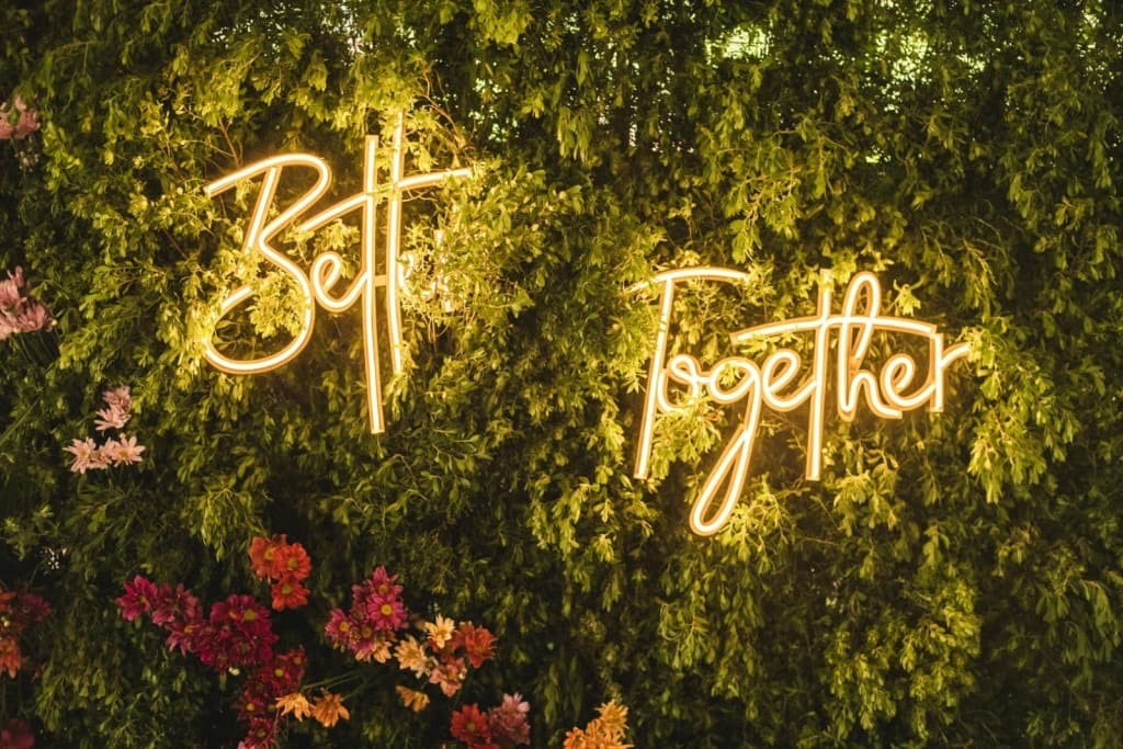 Un cartel de neón cálido que dice Better Together sobre un muro de follaje, con flores de colores montadas abajo.