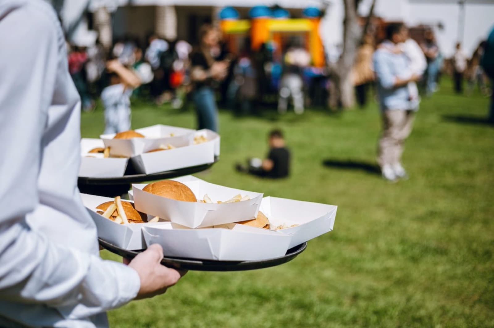 Dos bandejas de sándwiches en la mano de un mozo, con la fiesta al aire libre desenfocada detrás.