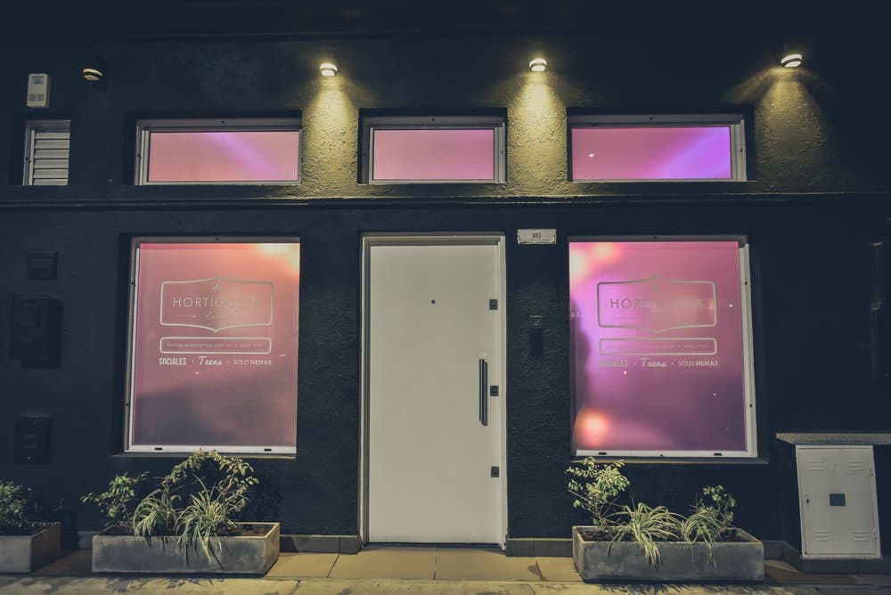El frente del salón desde la vereda, de noche: fachada gris oscuro con puerta blanca, ventanas iluminadas de rosa con el nombre del salón y macetas con plantas a los costados.