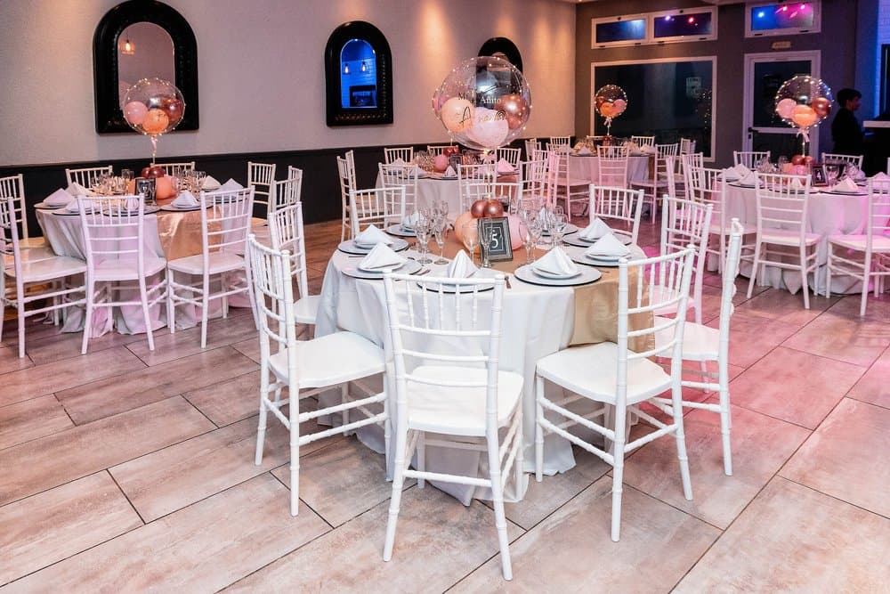 El salón con luz de día, con mesas redondas de mantel blanco y beige, sillas Chiavari blancas, globos rosados de centro de mesa y piso de madera clara.