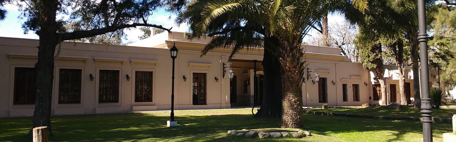 La casona blanca de una planta con sus ventanas de madera, faroles negros en la pared y la galería de entrada, vista desde el pasto del parque entre pinos y una palmera.
