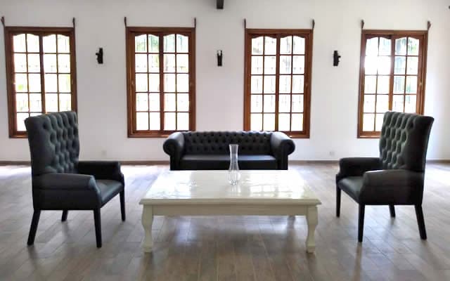Un salón vacío de paredes blancas y piso de madera clara, con cuatro ventanales de marco de madera y reja, un sillón capitoné de cuero oscuro y dos butacas altas alrededor de una mesa ratona blanca.