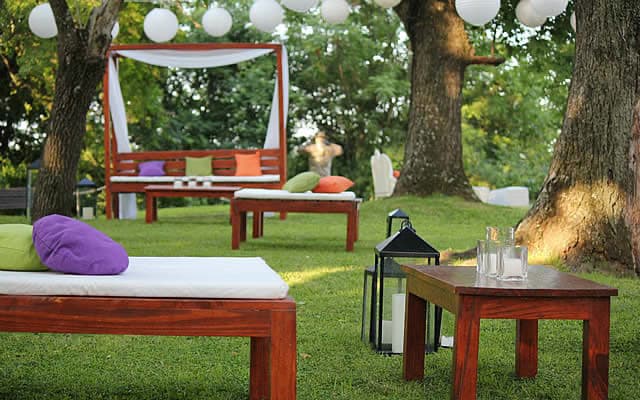 El sector de living al aire libre en el parque, con camastros de madera y almohadones verdes y naranjas, mesas bajas con faroles y velas, y esferas de papel blancas colgadas entre los árboles.