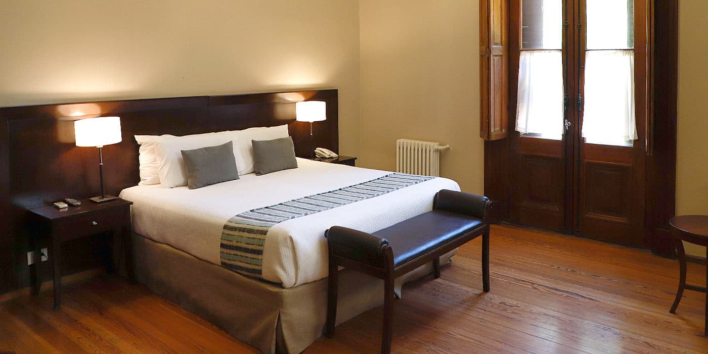 Una habitación del casco con cama king, piso de madera lustrada, banqueta de cuero a los pies y puertaventanas de madera oscura con cortinas blancas.