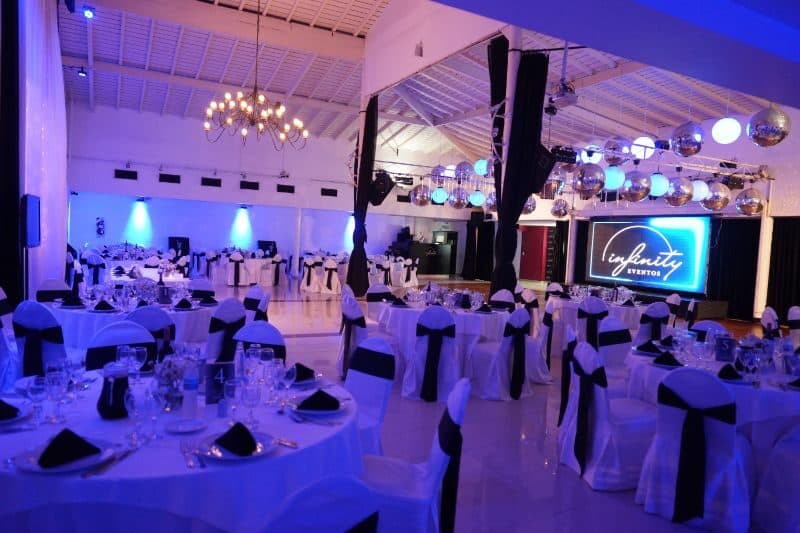 La sala grande en luz azul, con techo a dos aguas, una araña de caireles, mesas con sillas vestidas de blanco y moño negro, y una pantalla encendida al fondo.