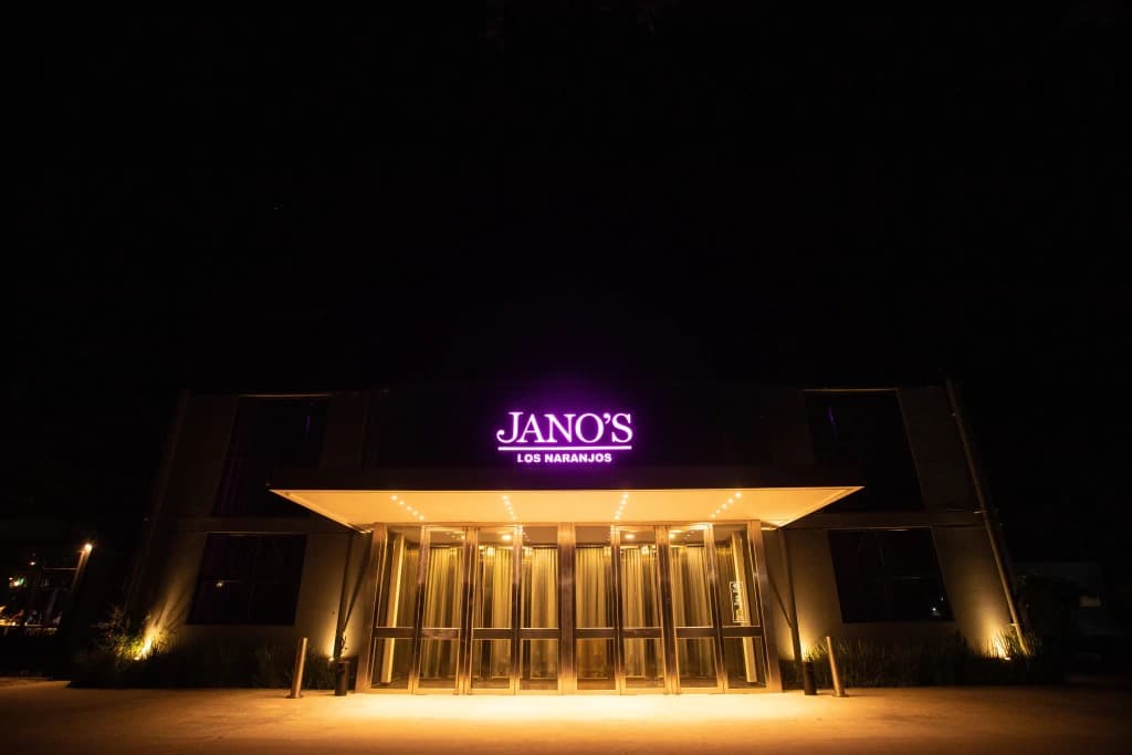 La fachada del salón de noche, con el cartel luminoso que dice Jano's Los Naranjos sobre la marquesina y las puertas vidriadas iluminadas desde adentro.