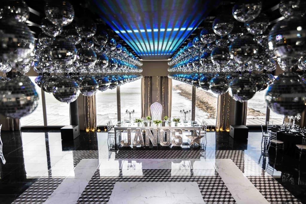 La pista de damero blanco y negro bajo un techo cargado de bolas de espejo, con la mesa principal y las letras luminosas contra el ventanal al mar.