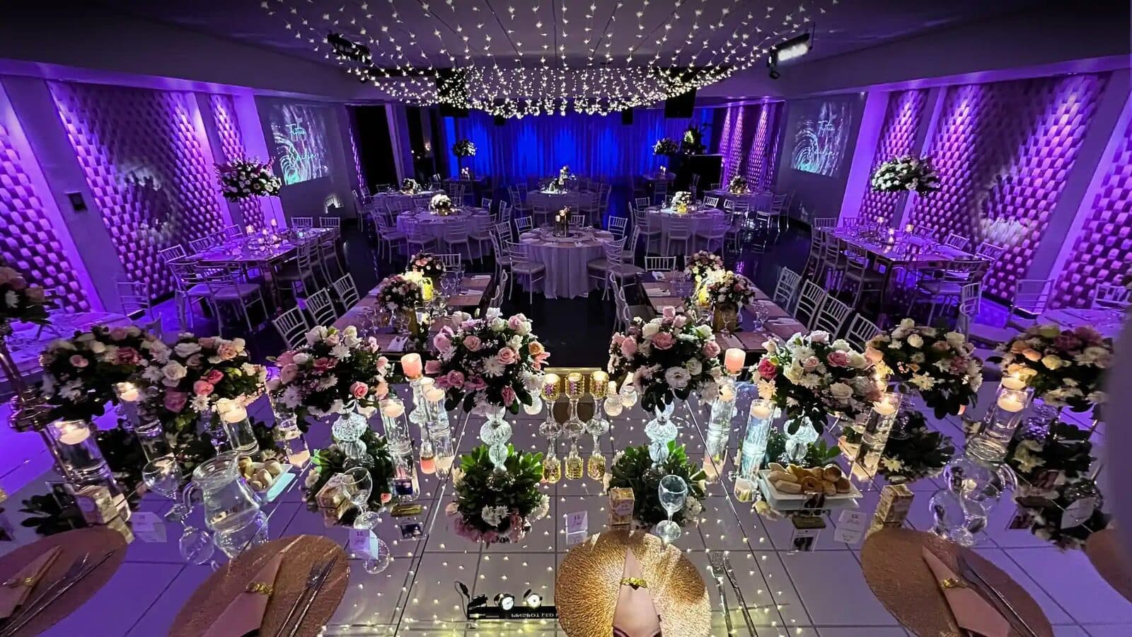 El salón con luz violeta y cortina de luces en el techo, una mesa larga en primer plano con centros de flores y cristal, y mesas redondas al fondo.