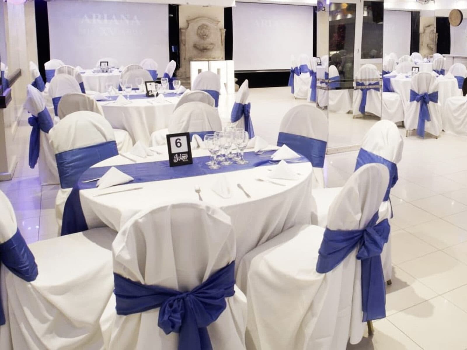 El salón armado para una fiesta de quince, con mesas redondas de mantel blanco, sillas vestidas con moños azules y una pantalla proyectada al fondo.