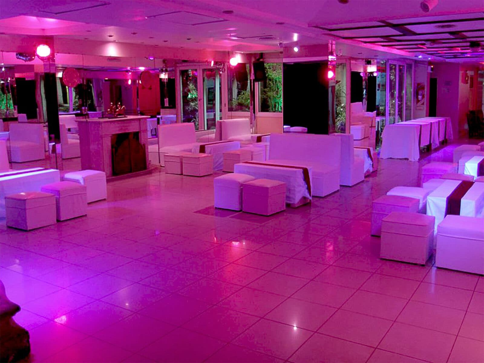 La pista de baile iluminada de rosa, rodeada de livings y puffs blancos, con la chimenea y los ventanales a la vegetación al fondo.