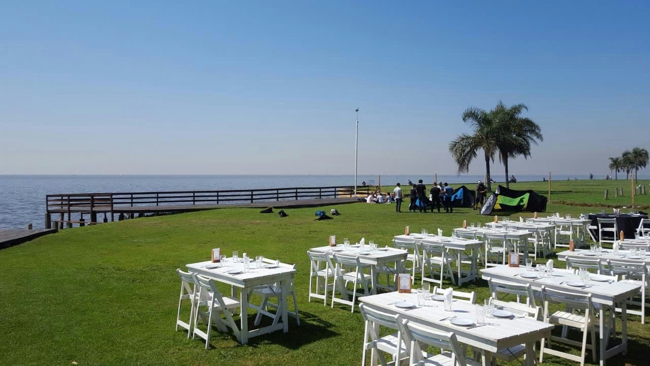 El predio con mesas y sillas plegables blancas puestas sobre el césped al borde del agua, un muelle de madera que se mete en el río a la izquierda y palmeras contra el cielo despejado.