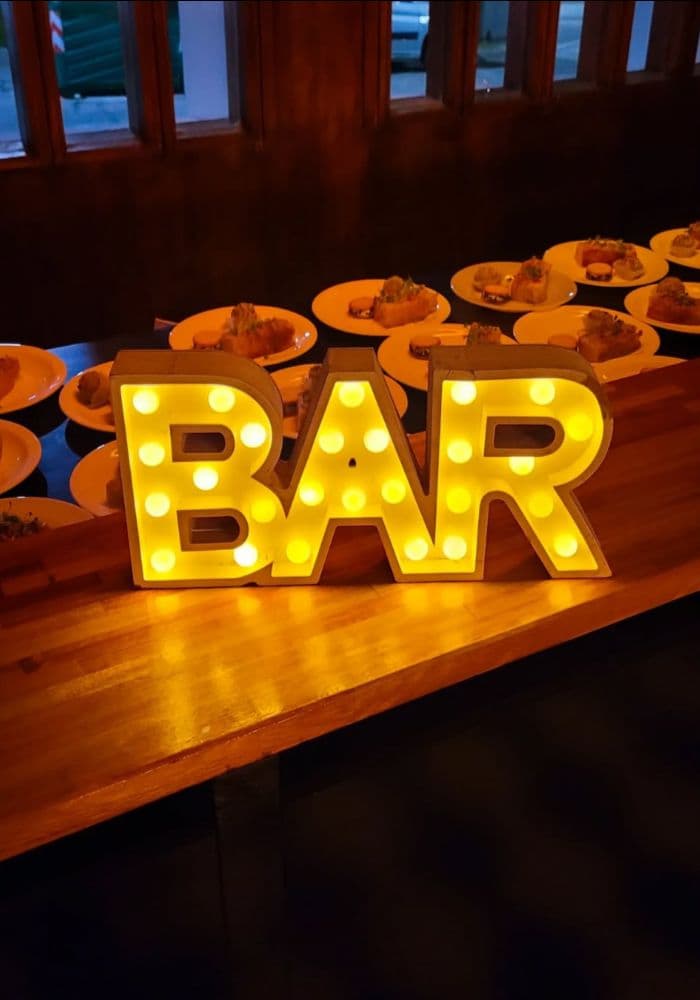 Las letras luminosas de BAR encendidas sobre la barra de madera, con las bandejas de bocados servidas detrás.