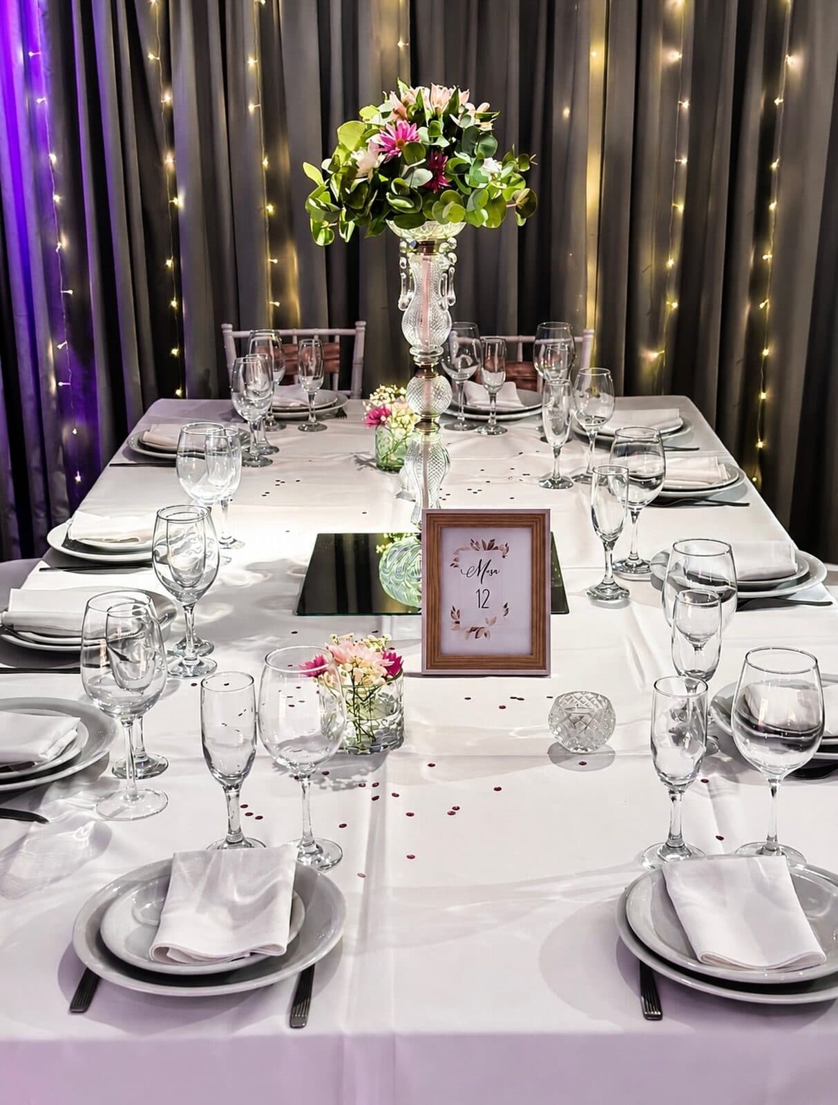 Mesa larga vestida de blanco con vajilla y copas, un centro de flores alto, pétalos sueltos sobre el mantel y una cortina de luces cálidas al fondo.