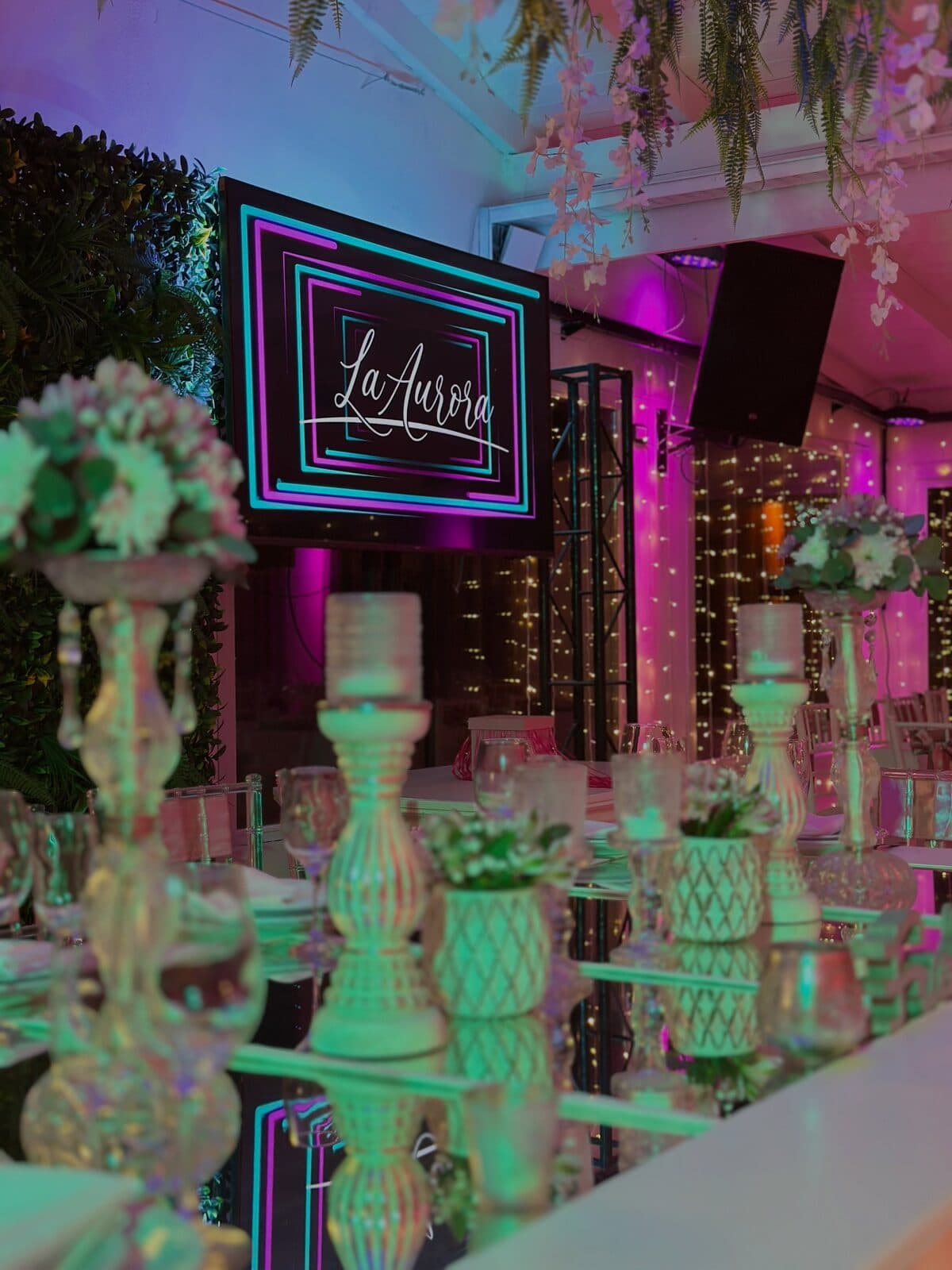 El salón con el cartel luminoso del lugar sobre una pared de follaje, candelabros y centros de flores sobre una mesa espejada, y luces violetas al fondo.