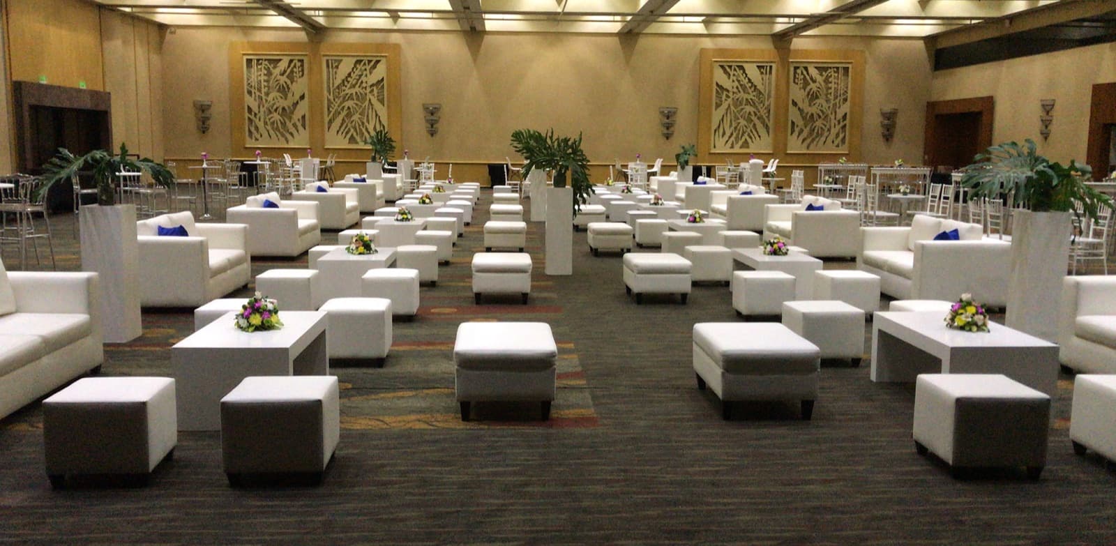 Un salón montado con los livings blancos y los puffs de la empresa, con mesas bajas y ramos chicos de flores encima.