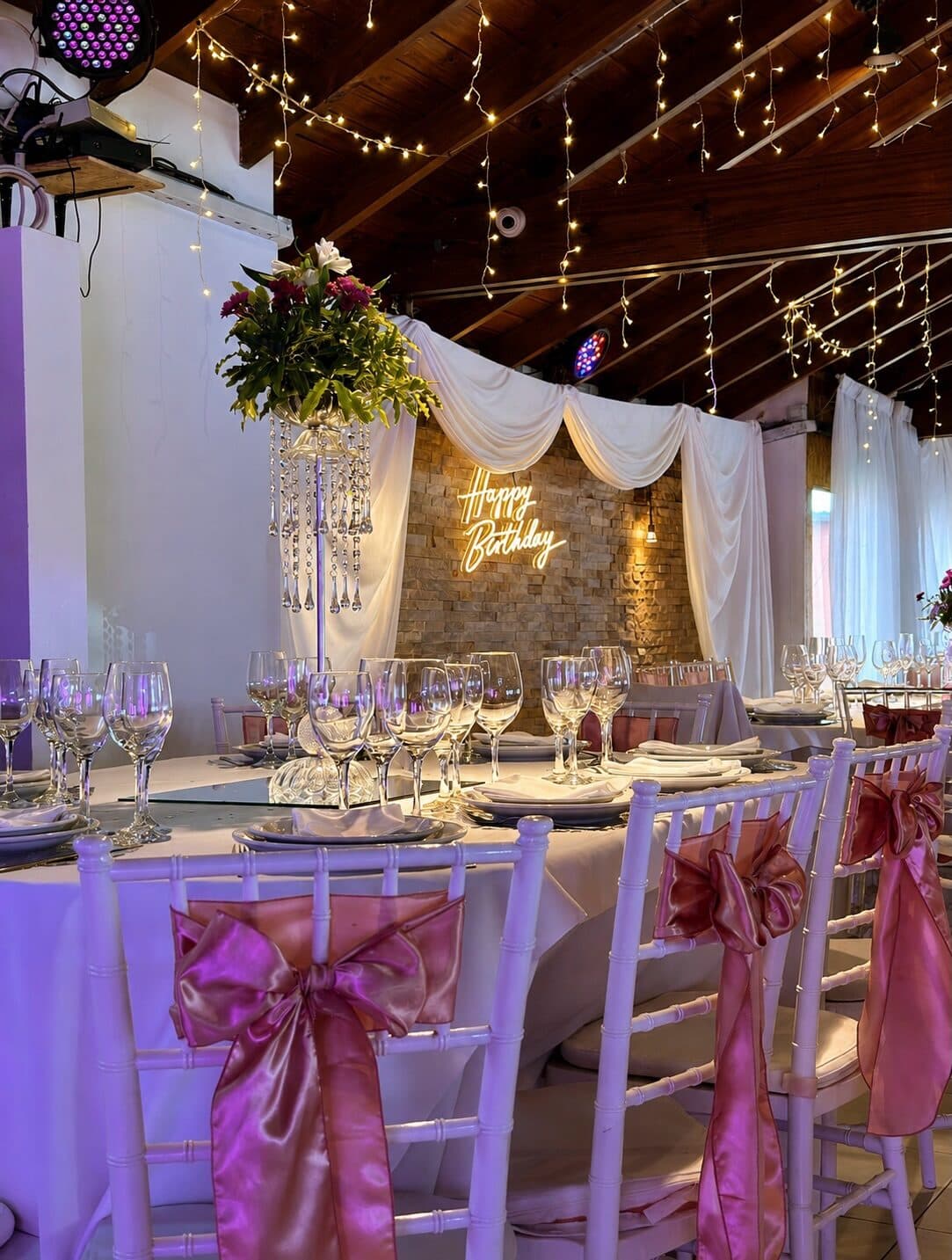 El salón armado con mesas de mantel blanco, sillas Chiavari con moños rosados, un centro de flores alto y un cortinado claro sobre la pared de piedra al fondo.